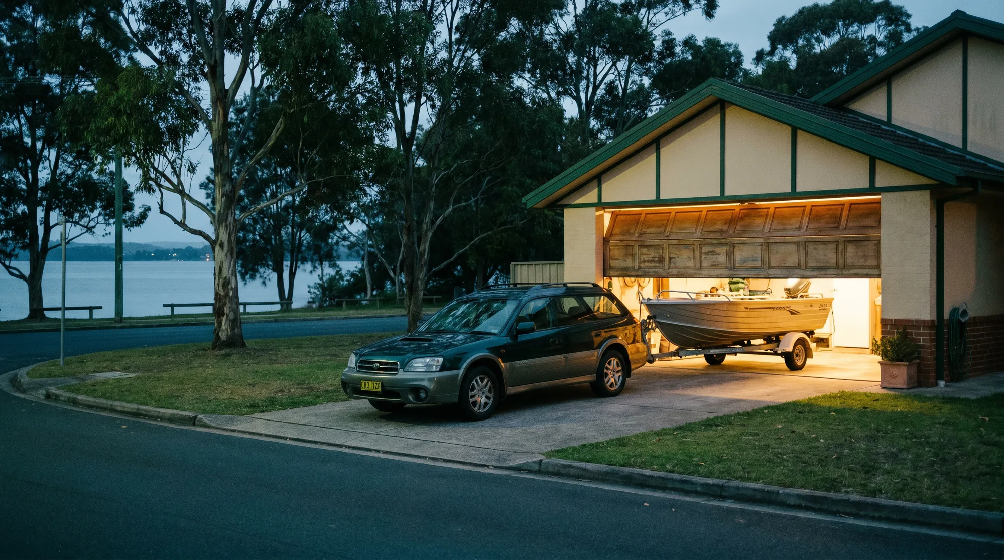 A garage door half open at first light, warm light spilling onto the driveway where a boat trailer waits hitched to a wagon